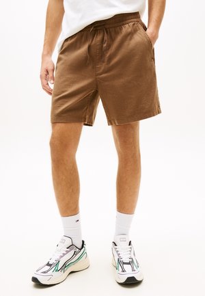 Άνδρας που φοράει καφέ σορτς με κορδόνι, λευκές κάλτσες μέχρι τον αστράγαλο και λευκά αθλητικά παπούτσια με μαύρες και πράσινες λεπτομέρειες, στέκεται μπροστά σε απλό φόντο.