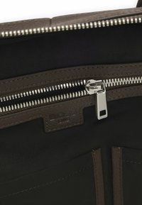 Intérieur d'un sac en cuir brun montrant une fermeture éclair argentée et une étiquette en relief "BOSS ONE" sur un patch en cuir cousu.