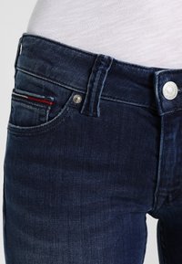 Jeans de mezclilla azul oscuro con un diseño ajustado, que presentan un cierre de botón metálico, costuras sutiles y un pequeño bolsillo con acentos en rojo y blanco.