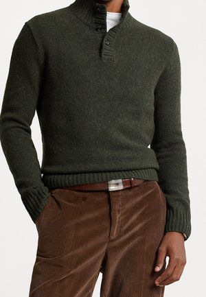 Homme portant un pull en maille vert foncé avec col boutonné, pantalon en velours côtelé marron, et ceinture en cuir marron avec une boucle argentée.
