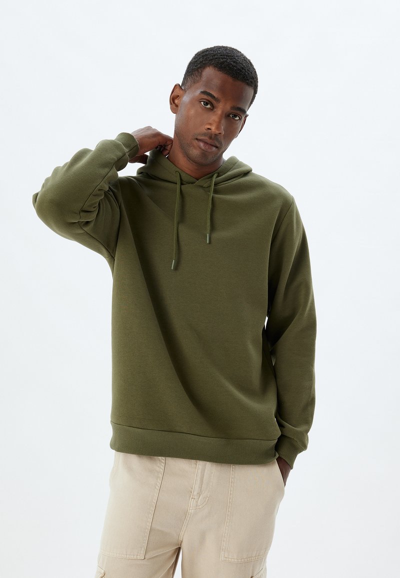 Sudadera de color verde oliva hecha de tejido texturizado; cuenta con capucha con cordón y puños acanalados. Llevada con pantalones cargo de colores claros.