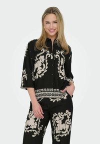 Chemise noire à boutons avec un motif floral crème, présentant une coupe décontractée, des manches trois-quarts et une bande texturée à l'ourlet.