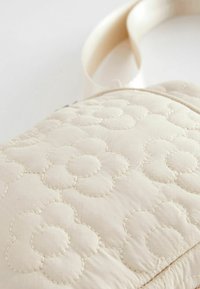 Kvistad beige axelväska med präglat blommönster, med en mjuk yta och en bred, mjuk rem. Dragkedjestängning upptill.