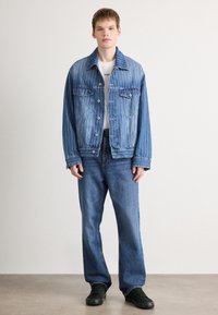 Τζάκετ από τζιν με κάθετες ρίγες, δύο τσεπάκια στο στήθος και μεταλλικά κουμπιά; συνδυασμένο με μπλε τζιν και μαύρα αθλητικά παπούτσια. Απλός, χαλαρός σχεδιασμός.