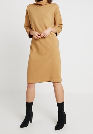 Vrouw draagt een knielange, geribbelde camelkleurige coltrui jurk met driekwartmouwen en zwarte enkellaarsjes met hakken, staand tegen een effen achtergrond.