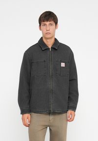 Črna jeans jakna z zadrgo na sprednji strani, dvema žepoma na prsih in ovratnikom. Značilnosti vključujejo vidno šivanje in majhno logotipno nalepko na levem žepu.