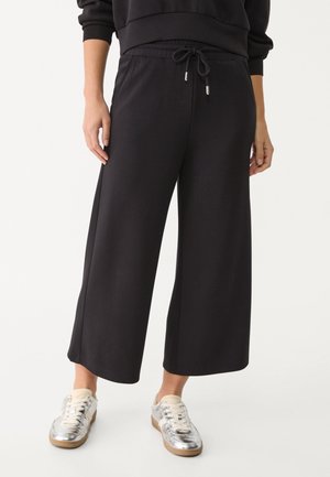 Zwarte wijde cropped broek met aantrekkoord in de taille, gedragen met zilverkleurige metallic sneakers op een staand persoon.