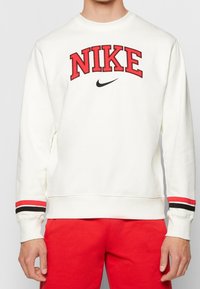 Vit sweatshirt med röda och svarta randiga muddar, "NIKE"-logotyp i röd med svarta accenter, gjord av mjukt tyg med rundad halsdesign.