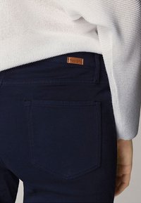 Donkerblauwe broek van katoen, met een achterzak en een leren merklabel in bruin, deels bedekt door een gebreide witte trui.