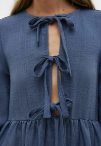 Temno moderna lanena obleka z okroglim izrezom, ki ima spredaj tri čvrsto zavezane pentlje in nabrano krilo. Teksturirana tkanina z naravnim videzom.