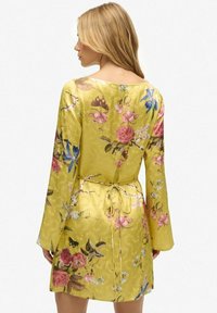 Geel bloemenjurk met lange mouwen, voorzien van een strik in de taille, satijnen textuur, en kleurrijke bloemen- en vlinderpatronen over het geheel.