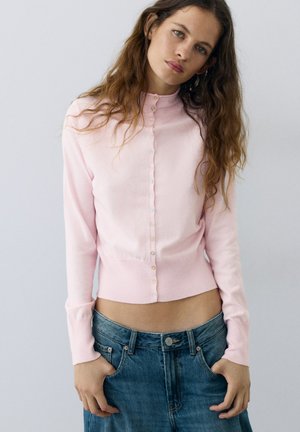 Femme aux longs cheveux ondulés portant un cardigan boutonné rose pâle et un jean bleu, debout les mains dans les poches, fond neutre.