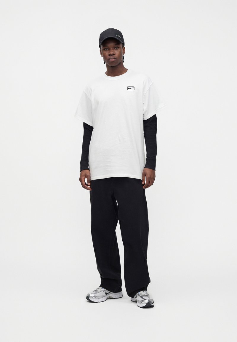 Junger Mann trägt schwarze Mütze, weißes kurzärmeliges T-Shirt über schwarzen Langarmshirts, schwarze Hose und silberne Sneakers und steht vor weißem Hintergrund.