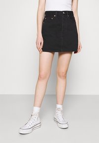 Minifalda de mezclilla negra con cintura alta, cierre frontal de botones y cinco bolsillos. Combinada con zapatillas altas blancas y calcetas.