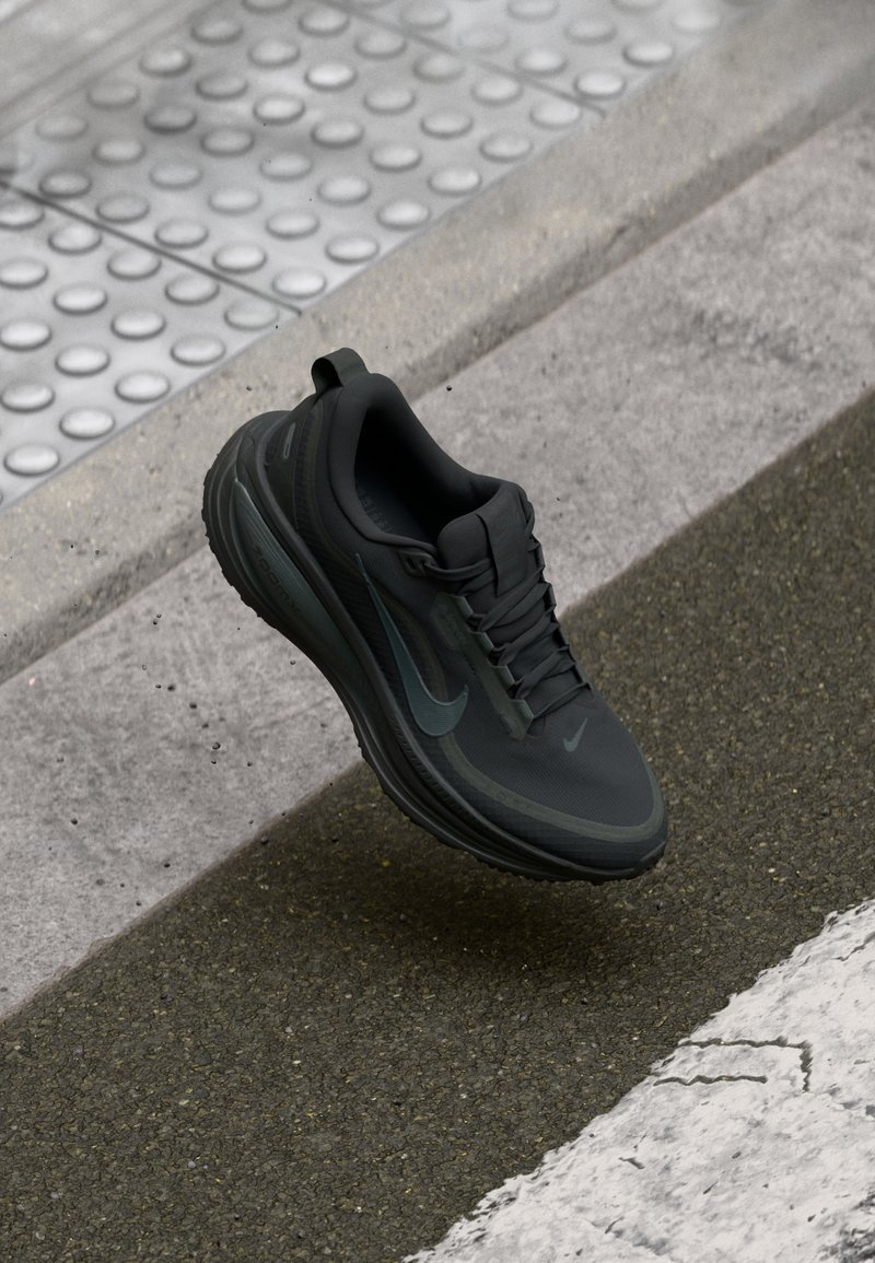 Black athletic shoe with smooth fabric upper, textured sole, and reflective accents. Features a rounded toe and lace-up design.