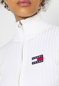 Pull blanc côtelé à fermeture éclair avec col montant et ferrures argentées. Présente un patch logo Tommy Jeans bleu, blanc et rouge sur la poitrine.