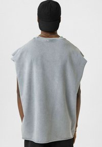 Grijze mouwloze shirt met een relaxte pasvorm, zachte textuur en een gebogen zoom. Draagt een zwarte pet en een zilveren ketting.