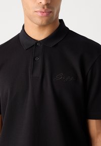 Črna polo majica iz teksturirane tkanine. Ima ovratnik, zapenjanje na dve gumbe in vezeno logo na levem prsnem delu.