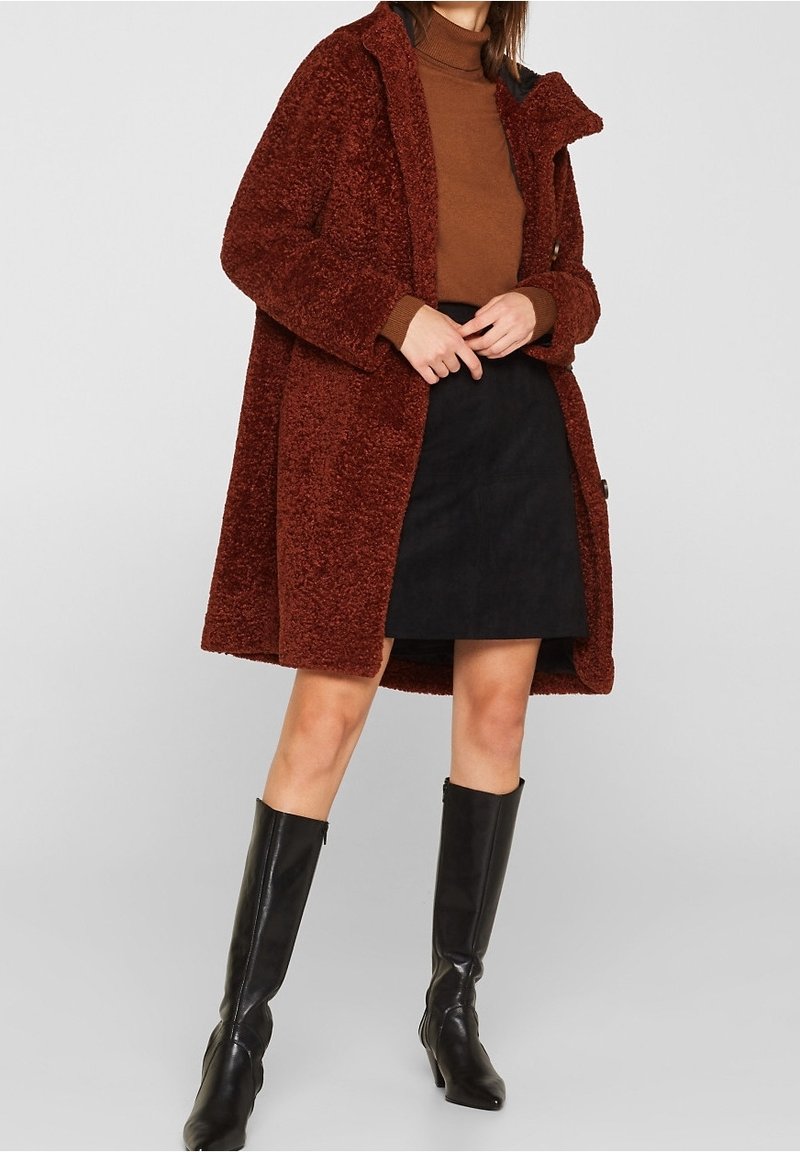 Femme portant un manteau texturé couleur rouille, un pull à col roulé marron, une jupe noire et des bottes noires montant jusqu'au genou sur un fond uni.