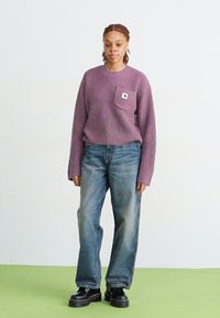 Suéter morado de canalé con un bolsillo en el pecho, combinado con jeans azules desgastados y botas negras gruesas, sobre un fondo claro y un suelo verde.