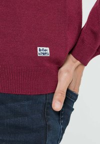 Pull en maille bordeaux avec des poignets et un bas côtelés. Présente une petite étiquette bleue et blanche avec le logo "Le Coq Sportif". Associé à un jean foncé.