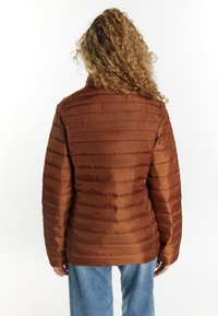 Brun, puffig jacka med sydda horisontella linjer, hög krage och slät, glänsande tyg. Bärs över ljusblå jeans.