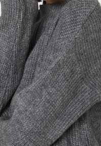 Personne portant un pull gris tricoté texturé à col montant, montrée des épaules jusqu'au milieu du torse sur un fond uni.