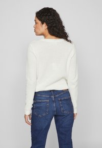 Pulover alb croșetat cu un model texturat, având decolteu rotund și mâneci lungi, purtat cu blugi din denim de culoare albastru închis.