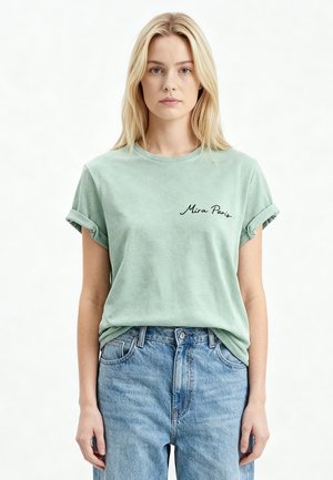 Femme aux cheveux blonds raides portant un t-shirt vert clair à manches retroussées avec le texte « Mira Paris » et un jean taille haute bleu clair.