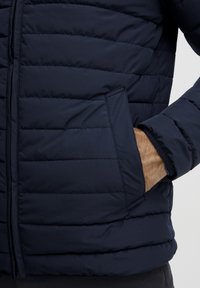 Jachetă puffer bleumarin, cu umplutură cusută orizontal, buzunar lateral și închidere cu fermoar; fabricată dintr-un material neted și ușor.