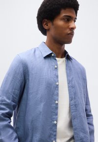 Νεαρός άνδρας με αφροκέντυ hairstyle, φοράει ένα ανοιχτό μπλε πουκάμισο πάνω από ένα λευκό T-shirt, κοιτάζοντας προς τα δεξιά σε απλό φόντο.