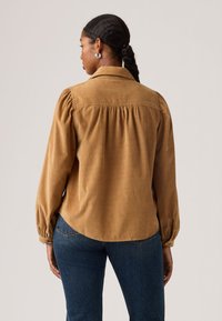 Brown corduroy shirt featuring a spread collar, long puffed sleeves, and gathered detailing at the back, styled with blue denim jeans.