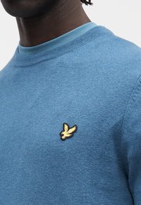 Modr pulover iz volne z rebrastim okroglim izrezom in majhnim črnim in zlato vezenim logotipom ptice na prsih.