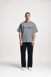 Camiseta de algodón gris con "2Y STUDIOS" impresa en negro, combinada con pantalones cargo negros y zapatillas blancas, de pie sobre un fondo neutro.