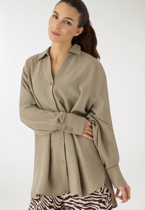 Donna con capelli lunghi e scuri che indossa una camicia beige oversize con bottoni e pantaloni a stampa zebra, in piedi contro uno sfondo bianco uniforme.