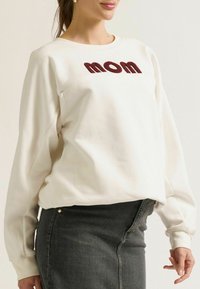 Lichtbeige sweatshirt met "mom" in bordeaux, ronde halslijn, lange mouwen en een relaxte fit, gecombineerd met een donkergrijze rok.