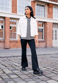 Valkoinen denim-takki mustavalkoisen raidallisen paidan päällä, yhdistettynä leveälahkeisiin mustiin housuihin ja tukeviiin mustiin kenkiin, tiiliseinän taustalla.