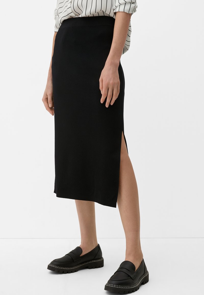 Schwarzer knielanger Rock mit einem seitlichen Schlitz, glatter Textur und tailliertem Design. Kombiniert mit schwarzen Slippern und einer gestreiften Bluse.