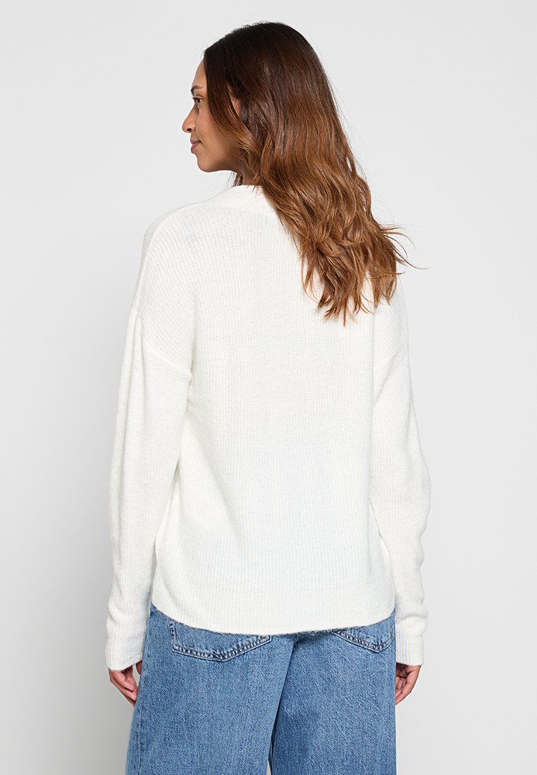Maglione bianco a maglia con vestibilità rilassata, texture a coste e spalle leggermente cadenti, abbinato a jeans a gamba larga di un azzurro chiaro.