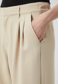 Beige nadrág, pliszézéssel és oldalsó zsebekkel. Simább anyagból készült, testreszabott derékkal, ami strukturált sziluettet ad.