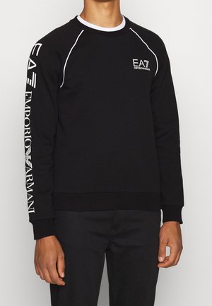 Czarna bluza z długim rękawem z białym przeszyciem, logo "EA7 Emporio Armani" na piersi oraz duże oznaczenie na lewym rękawie, noszona przez osobę.
