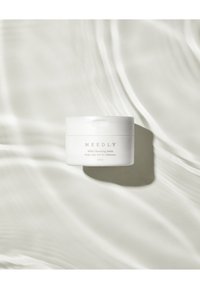 Un pot rond blanc de "NEEDLY Mild Cleansing Balm", étiqueté avec le nom du produit et le volume (120 ml), posé sur un fond lumineux réfléchissant.