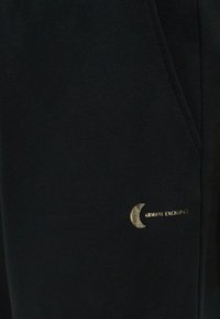 Melna auduma gabals ar mazu zelta pusmēness zīmi un tekstu "ARMANI EXCHANGE" tuvu šuvē vai kabatas malai.