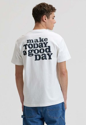 Junger Mann von hinten, trägt weißes T-Shirt mit fettem Schriftzug "make TODAY a good DAY" und blaue Jeans vor einfachem Hintergrund.