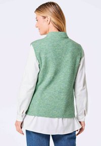 Groene, gemêleerde gebreide vest met een hoge kraag, gedragen over een witte blouse met lange mouwen, gecombineerd met blauwe denim jeans.