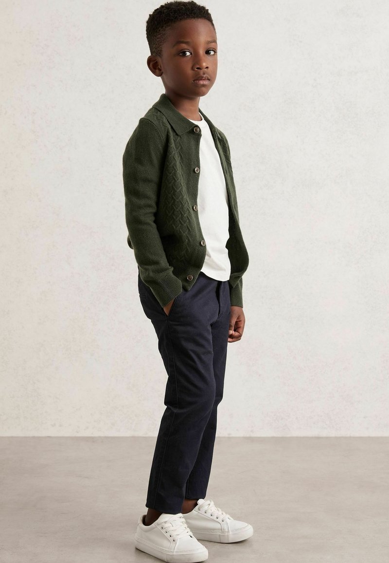 Junger Junge, der mit den Händen in den Taschen steht, trägt einen grünen Cardigan, ein weißes T-Shirt, dunkle Hosen und weiße Turnschuhe vor einem einfarbigen Hintergrund.