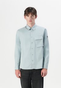 Camisa azul clara de mangas longas com colarinho abotoado, um bolso no peito e um patch de logotipo na manga. Tecido suave e design ajustado.
