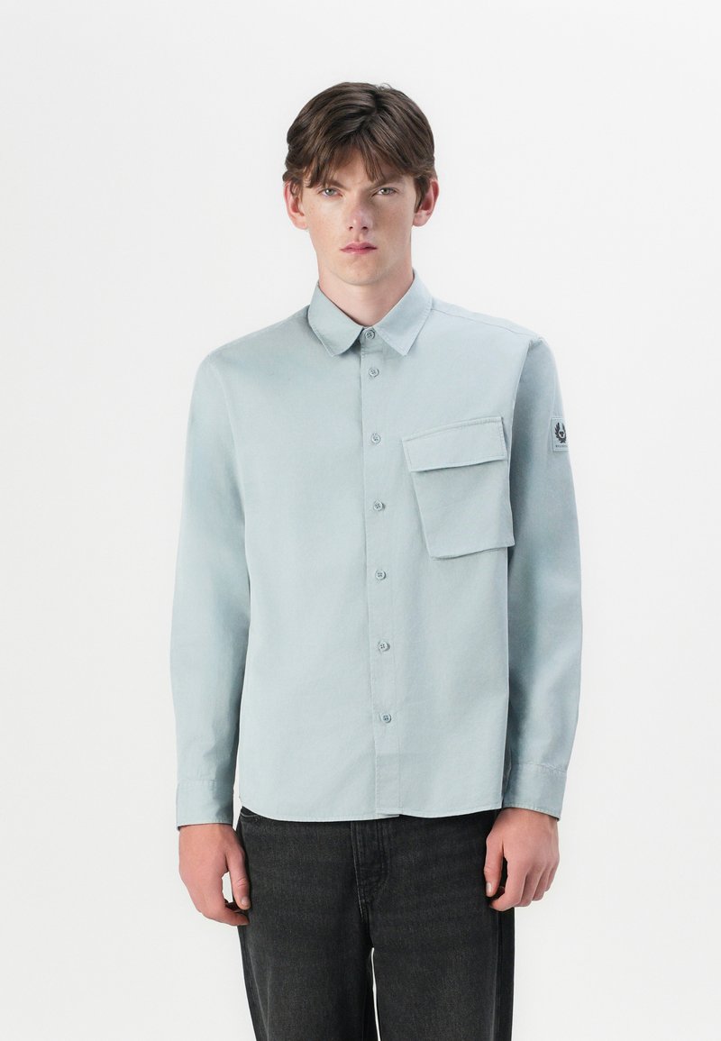Camisa azul clara de mangas longas com colarinho abotoado, um bolso no peito e um patch de logotipo na manga. Tecido suave e design ajustado.