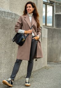 Lange bruine jas over een lichte trui, grijze slim-fit broek, en zwart-tan sneakers, gecombineerd met een kleine zwarte handtas. Soepele texturen, klassieke ontwerpen.