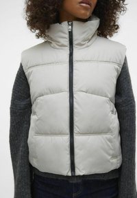 Gilet imbottito grigio chiaro con alto colletto e zip frontale, abbinato a un maglione scuro a trama. Presenta un motivo a righe orizzontali.